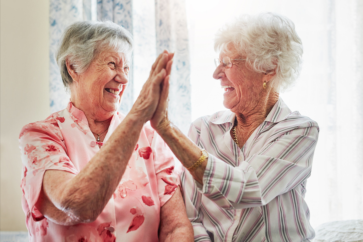 Two elderly women smiling and giving a high-five while sitting together in a bright room.