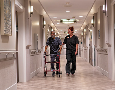 Caregiver assisting a senior man with a walker in a brightly lit hallway of a living community.