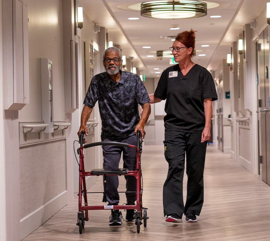 A caregiver assists an elderly man using a walker in a hallway of a senior living community.