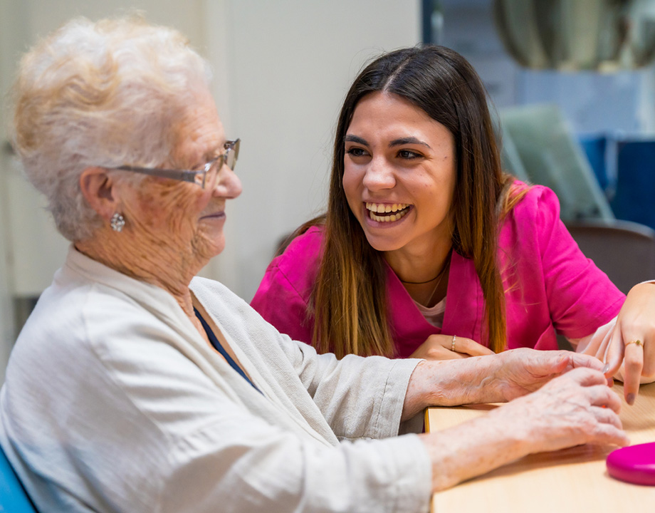 A young caregiver smiling and interacting with an elderly woman at a senior living community.