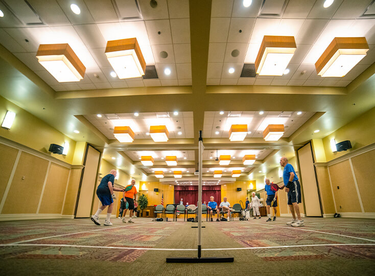 Residents playing indoor pickleball in a brightly lit community room at a senior living community.