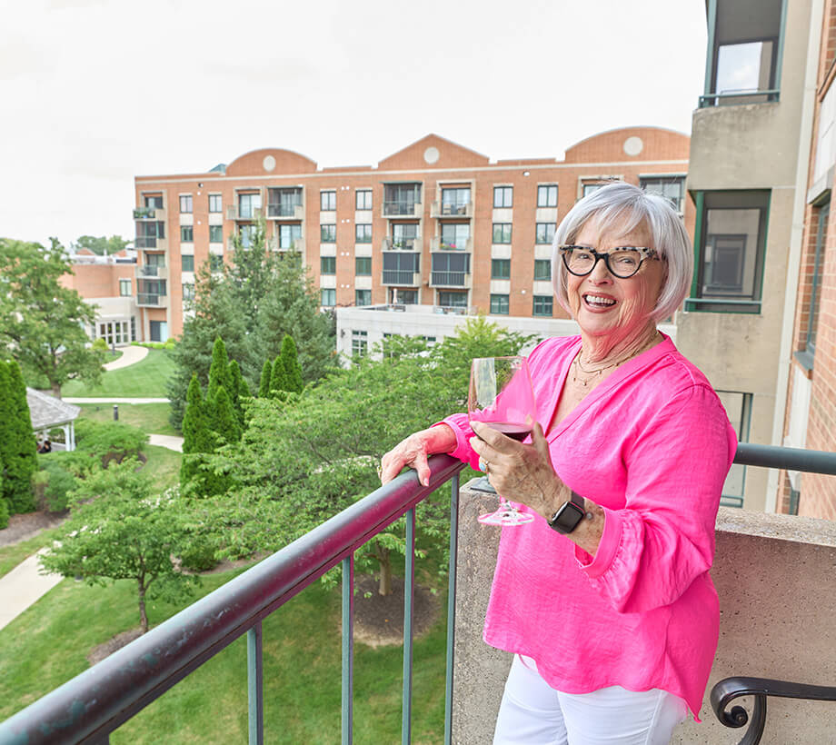 Elderly woman in a pink shirt enjoying a drink on a balcony overlooking a senior living community.