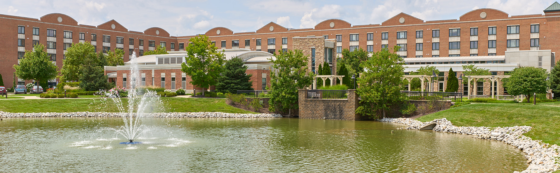 Elegant brick building with multiple units, greenery, and a fountain pond in front.