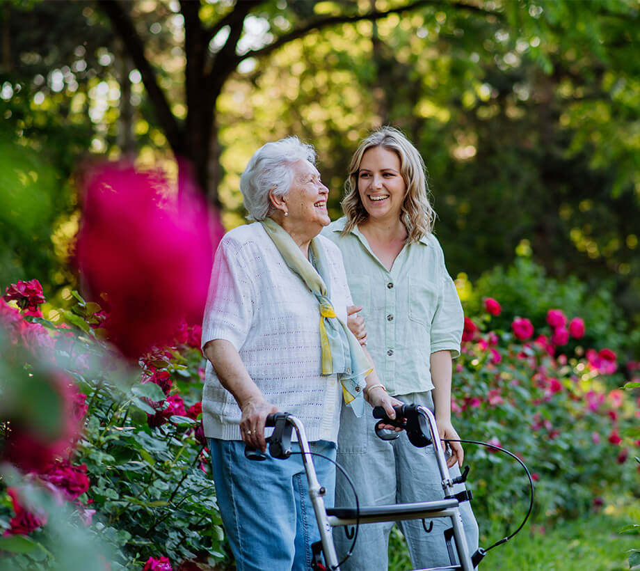 Senior woman using a walker enjoys a garden walk with a younger woman at a senior living community.