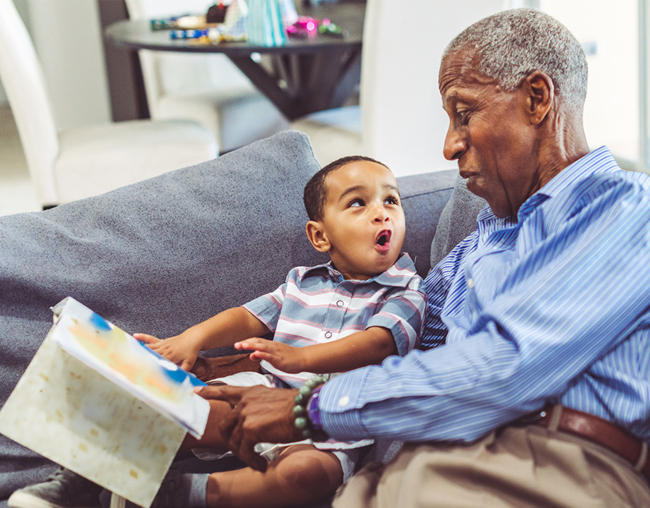 Elderly man in conversation with a young boy holding a book on a couch within living space.