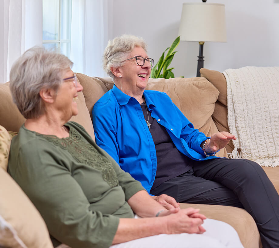 Two senior women laughing while sitting on a couch inside a cozy senior living unit.