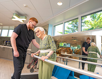 Elderly woman receiving physical therapy with support from a caregiver in a well-lit room.