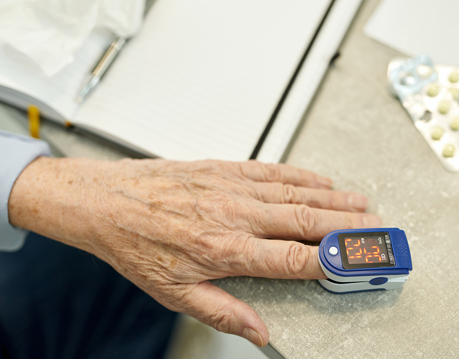 Elderly hand with pulse oximeter on finger next to pills and notepad on a table.