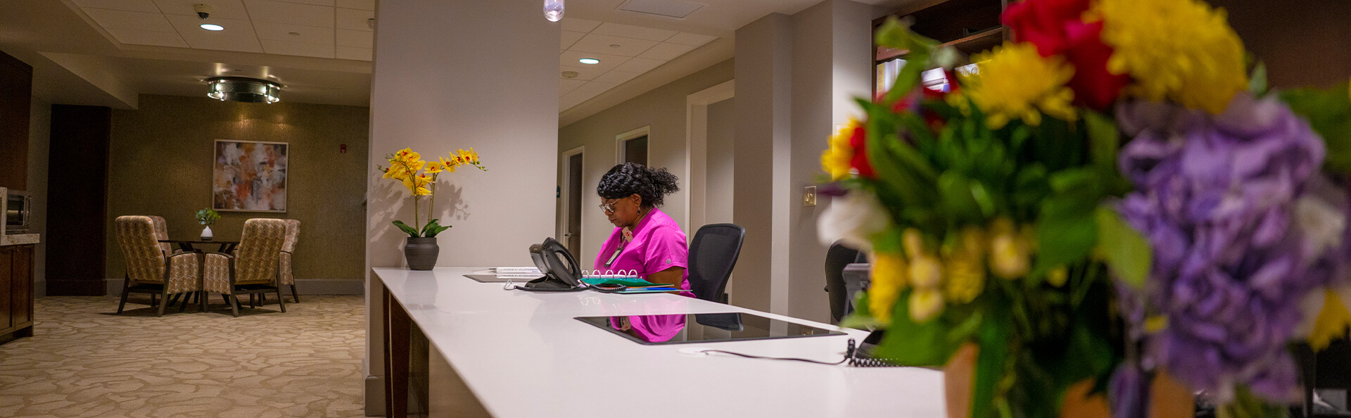 Reception desk with colorful flowers and staff, seating area in background.