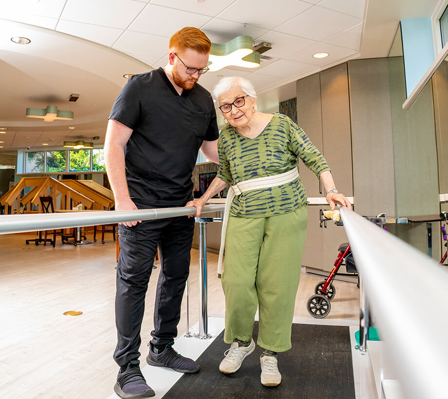 Physical therapist guiding elderly woman through rehabilitation exercises using parallel bars.