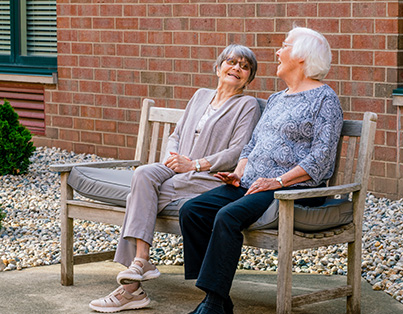 Two elderly women sitting and laughing on a bench outside a senior living community building.