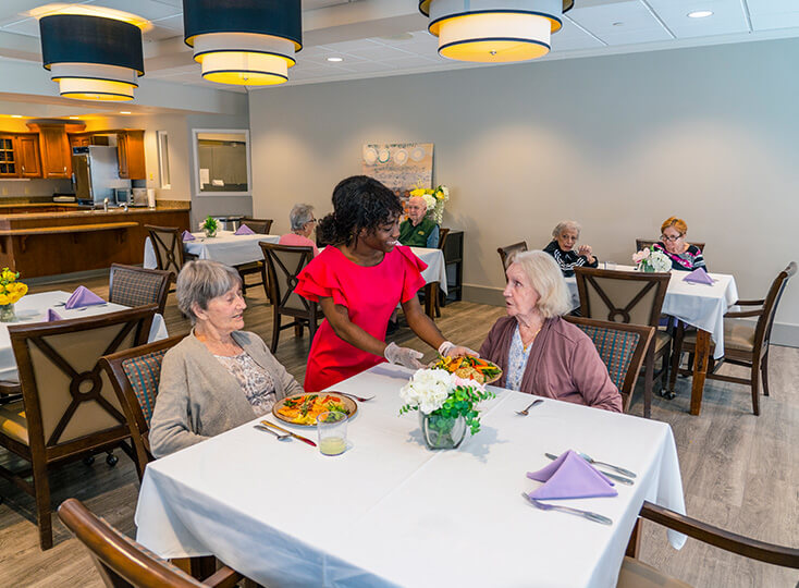 Dining area in a senior community with residents being served meals by a staff member.