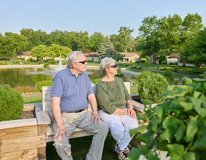 Two elderly people sitting on a bench in a garden at a senior living community, with a pond nearby.