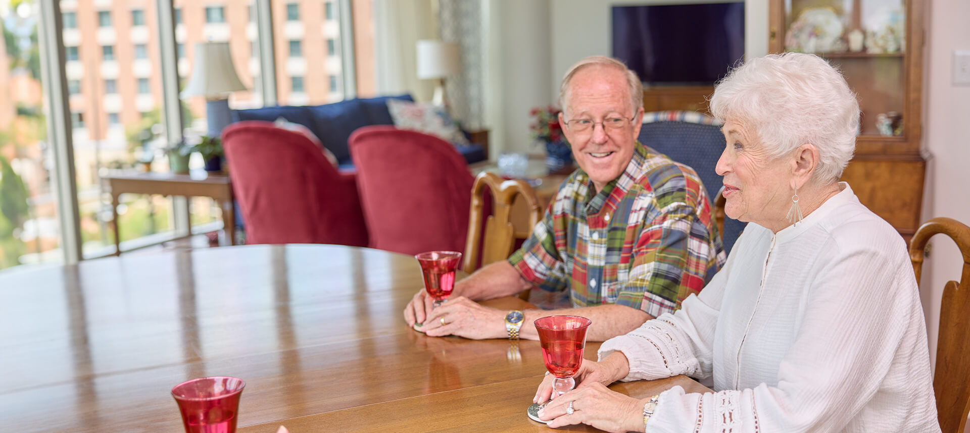 Two senior residents conversing at a dining table with red glasses in a bright room.