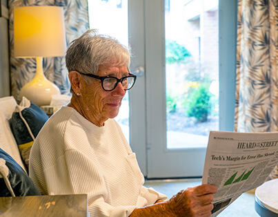 Senior woman reading a newspaper in a units living area with natural light and cozy decor.