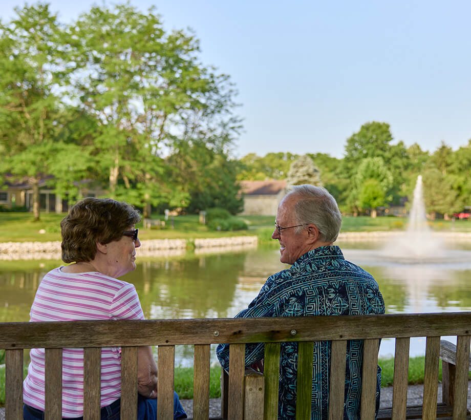 Two seniors sit on a bench by a pond with a fountain, surrounded by trees and greenery.