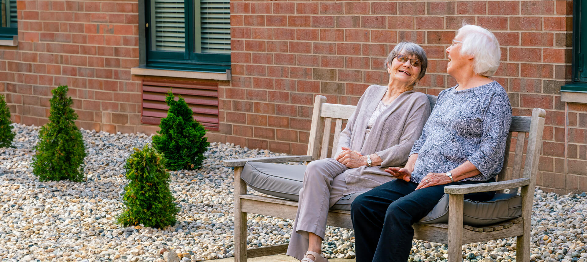 Two seniors sitting on a bench outdoors, laughing and enjoying their time outside a building.