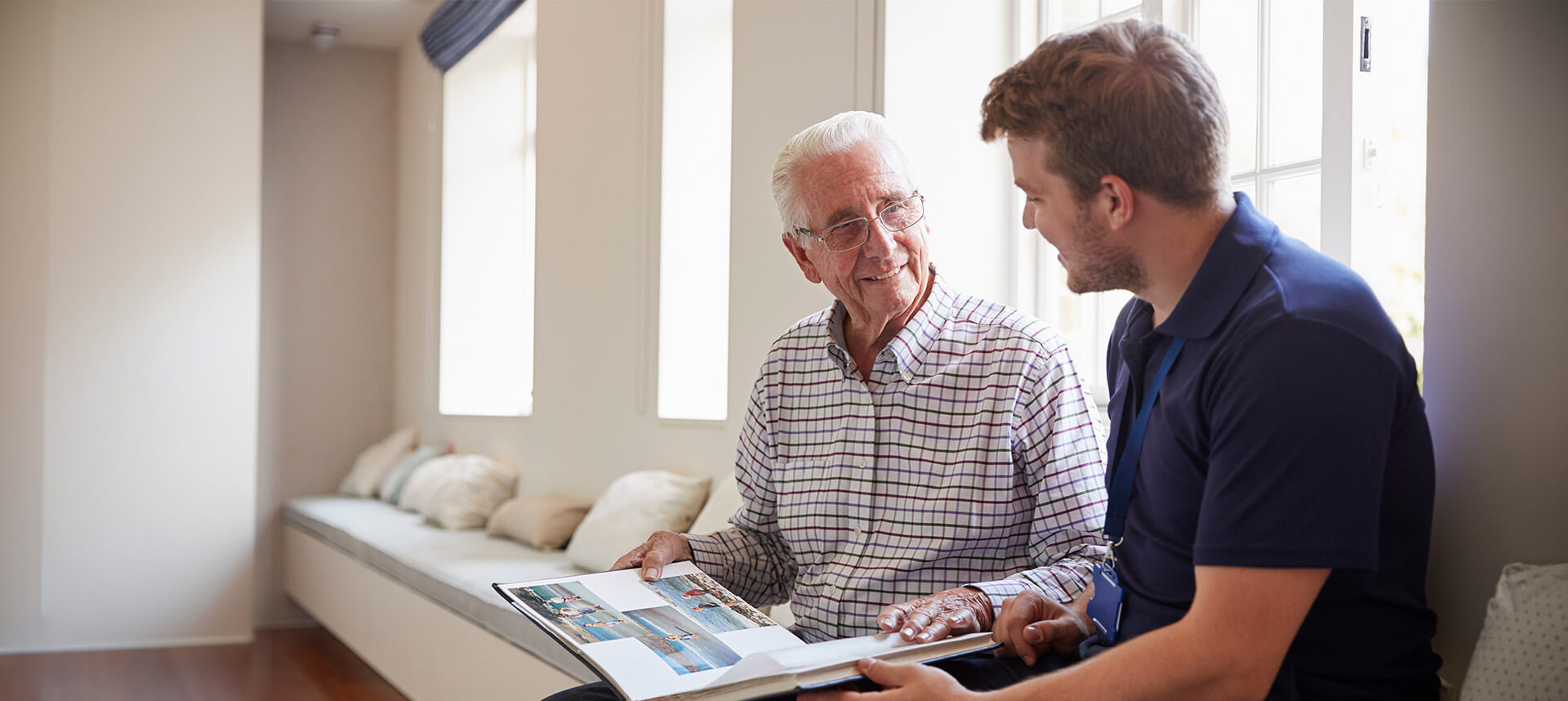 Older man showing a younger man a photo album while sitting by a window in a cozy room.