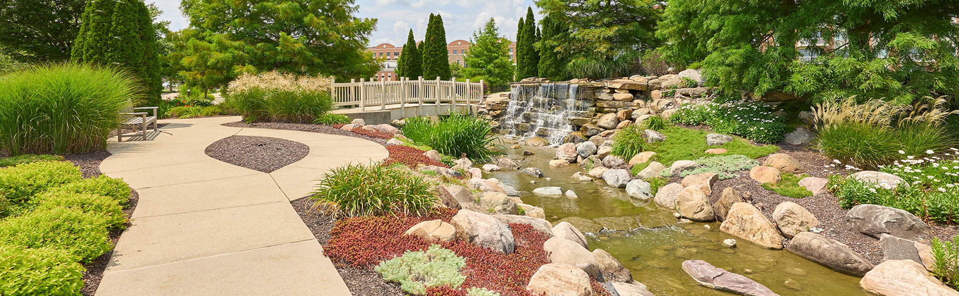 Serene garden with footpath, bench, small stone bridge, and cascading waterfall in senior community.