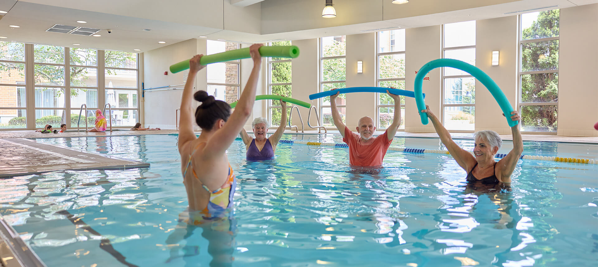 Residents in a pool at a senior living community engaging in a water aerobics class with pool noodles.